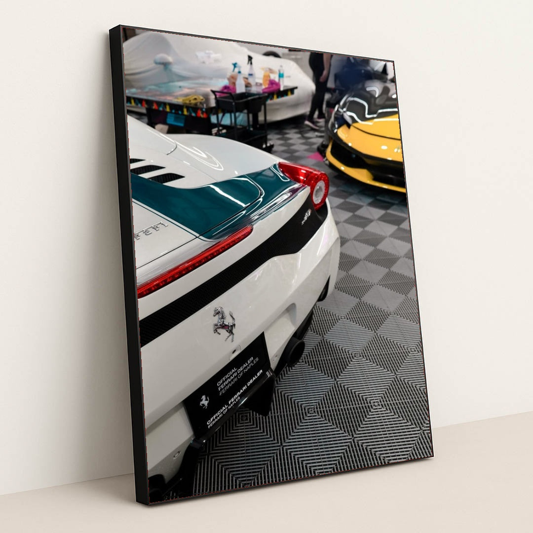 This photo shows the rear of a white Ferrari with a green stripe and a yellow Ferrari visible in an indoor detailing garage, in a black frame.