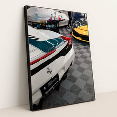 This photo shows the rear of a white Ferrari with a green stripe and a yellow Ferrari visible in an indoor detailing garage, in a black frame.