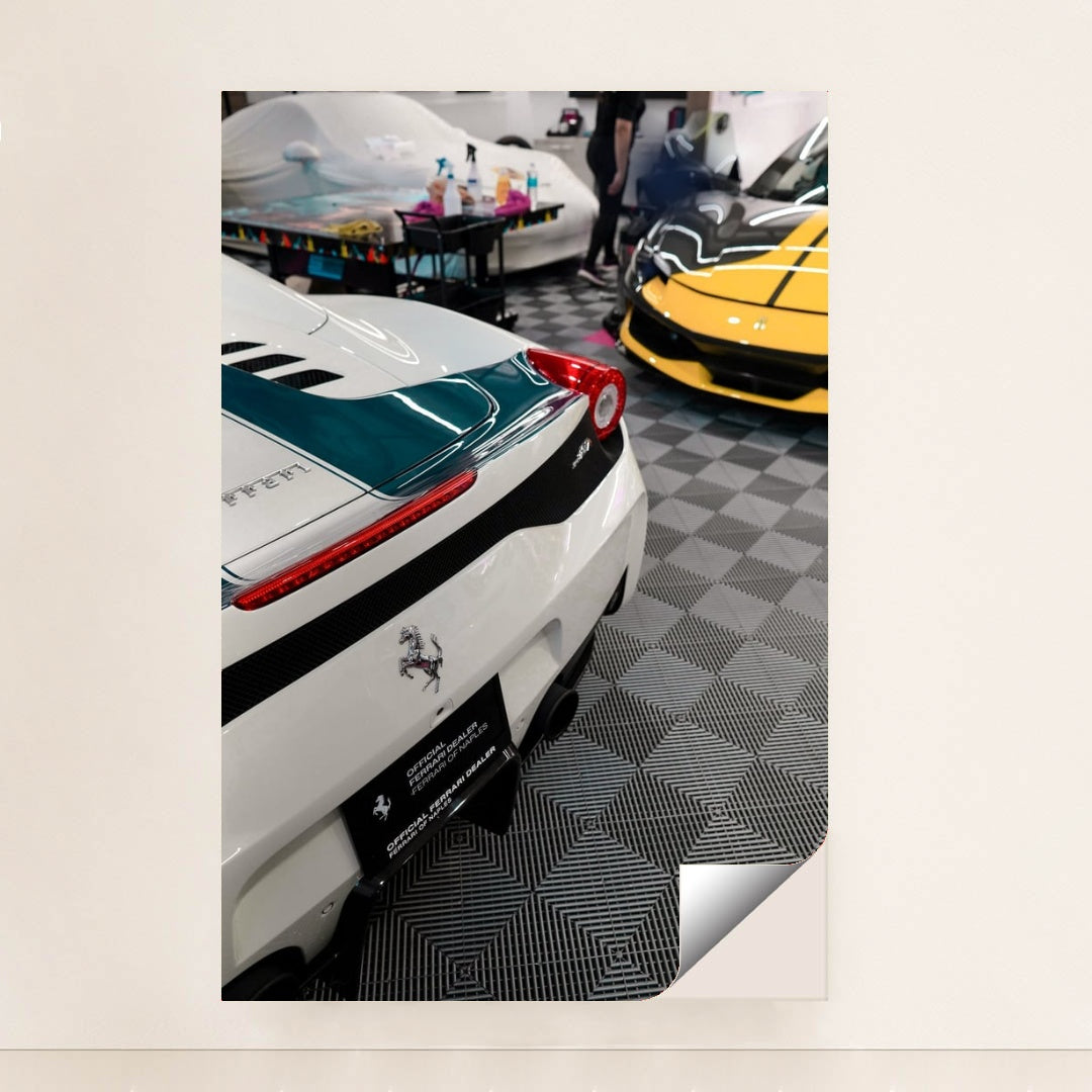 This photo shows the rear of a white Ferrari with a green stripe and a yellow Ferrari visible in an indoor detailing garage, unframed.