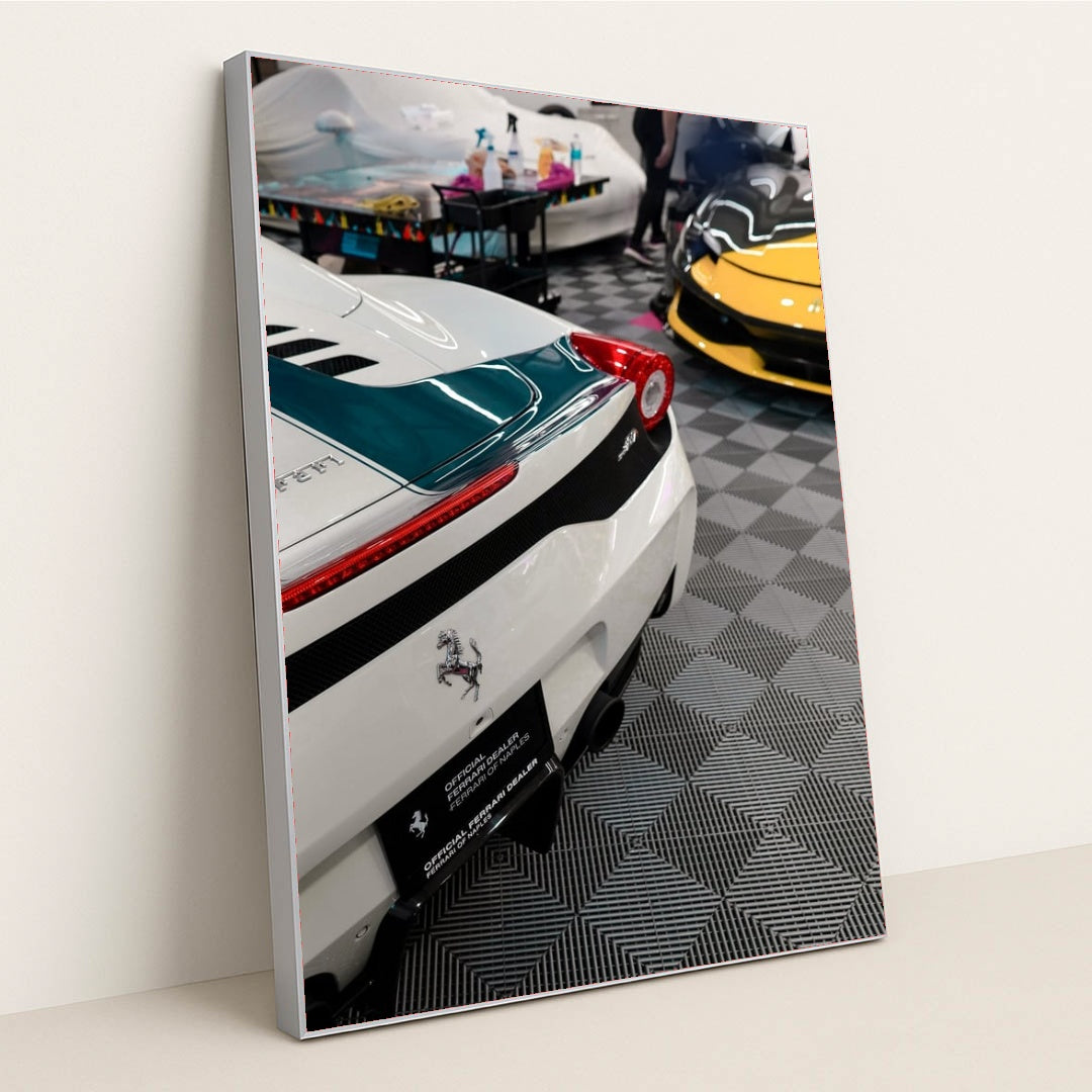 This photo shows the rear of a white Ferrari with a green stripe and a yellow Ferrari visible in an indoor detailing garage, in a silver frame.