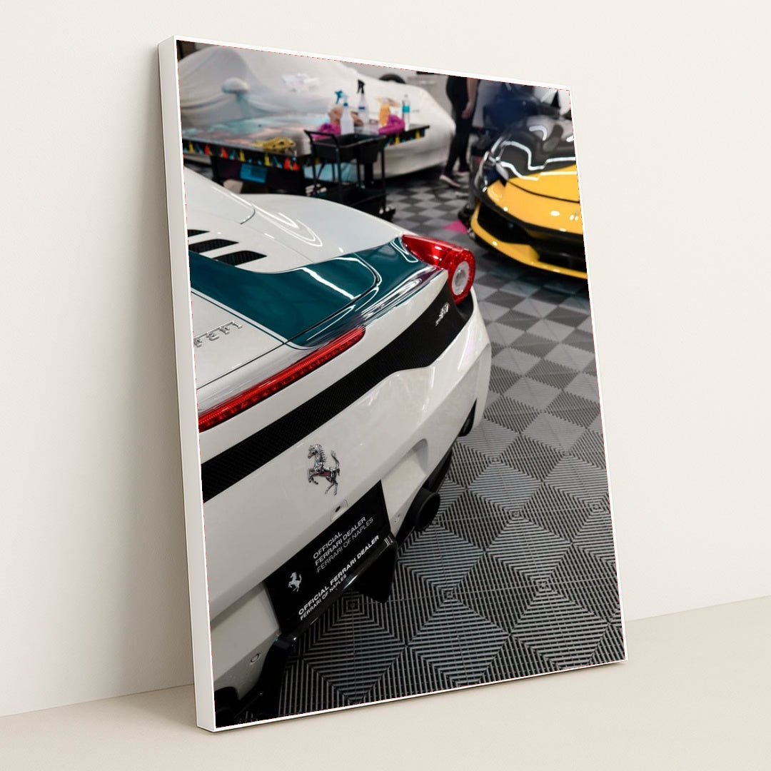 This photo shows the rear of a white Ferrari with a green stripe and a yellow Ferrari visible in an indoor detailing garage, in a white frame.