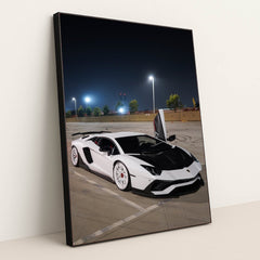 This photo shows a white Lamborghini Aventador with white wheels and an open door parked under streetlights at night, in a black frame.