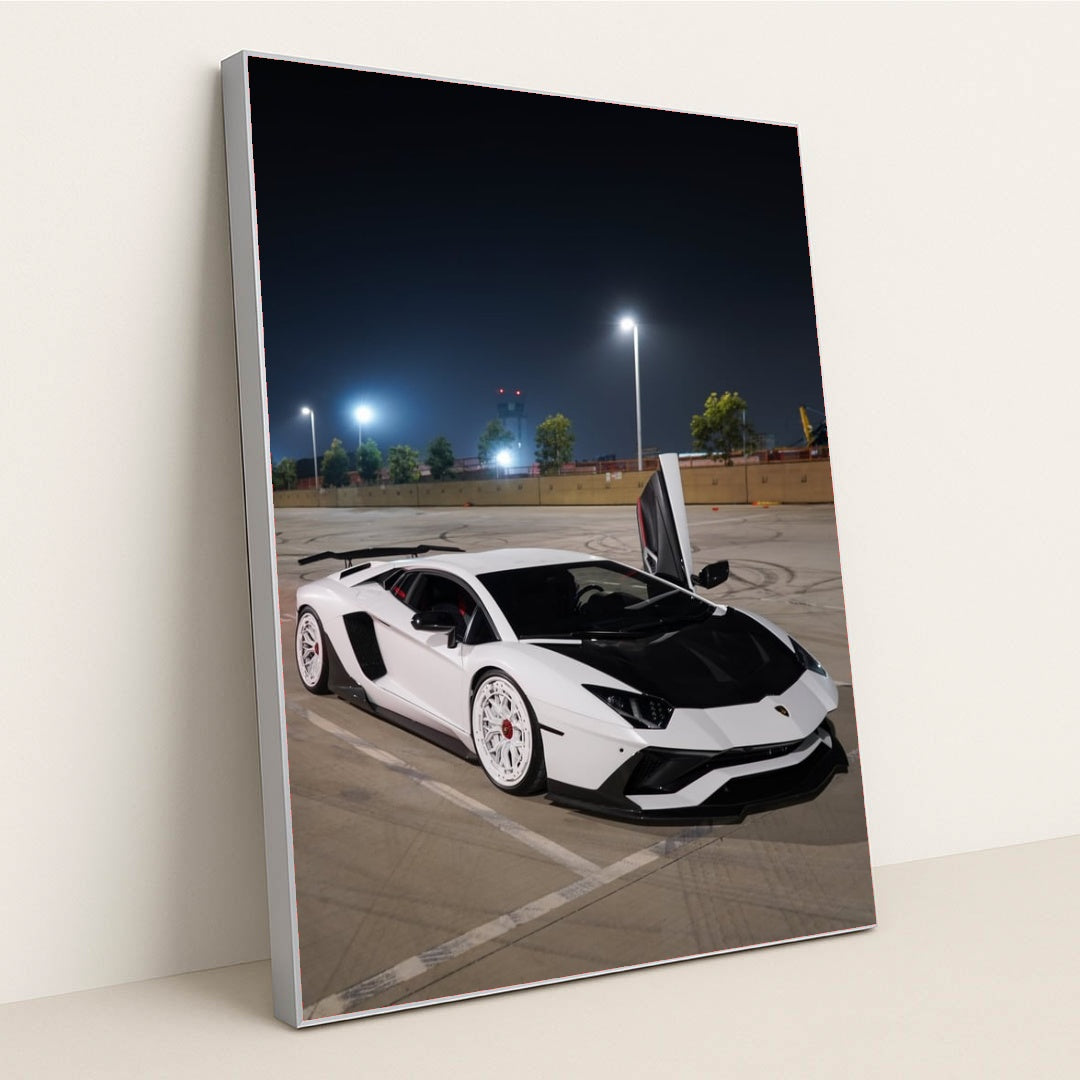 This photo shows a white Lamborghini Aventador with white wheels and an open door parked under streetlights at night, in a silver frame.