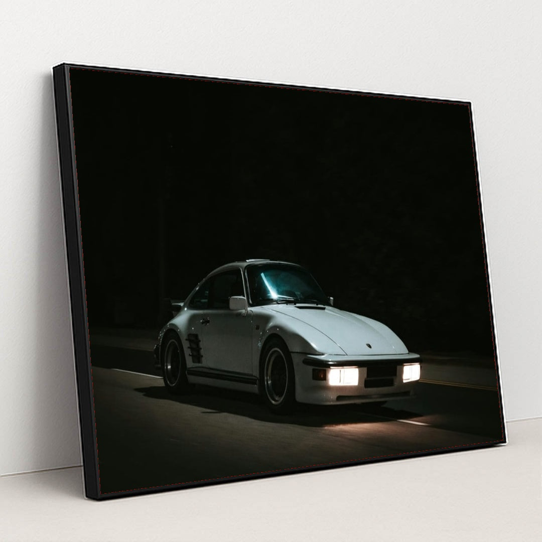 This photo shows a white Porsche 911 with pop-up headlights and wide body kit driving at night, in a black frame.