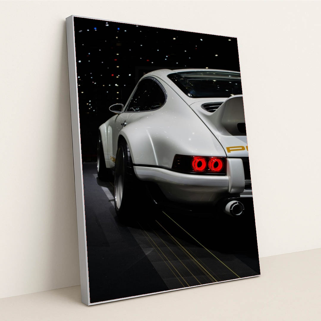 This photo shows the rear of a white Porsche sports car with a large wing, illuminated taillights, and starry reflections, in a silver frame.