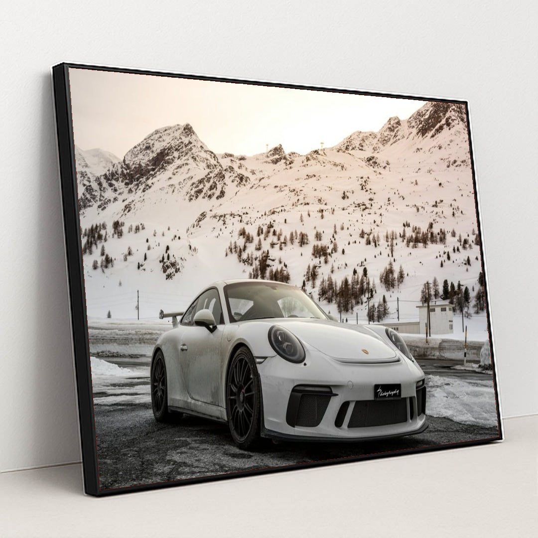 This photo shows a white Porsche sports car parked in front of snowy mountains at sunset, in a black frame.