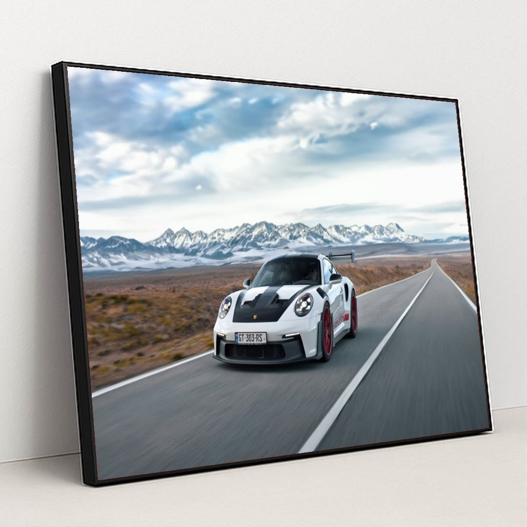This photo shows a white Porsche 911 GT3 RS with red wheels driving quickly on an open desert highway with mountains in the background, in a black frame.