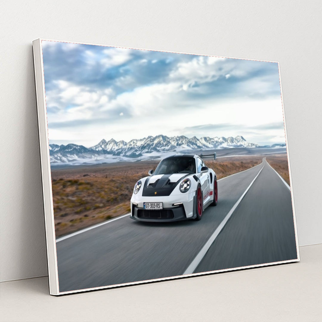 This photo shows a white Porsche 911 GT3 RS with red wheels driving quickly on an open desert highway with mountains in the background, in a white frame.