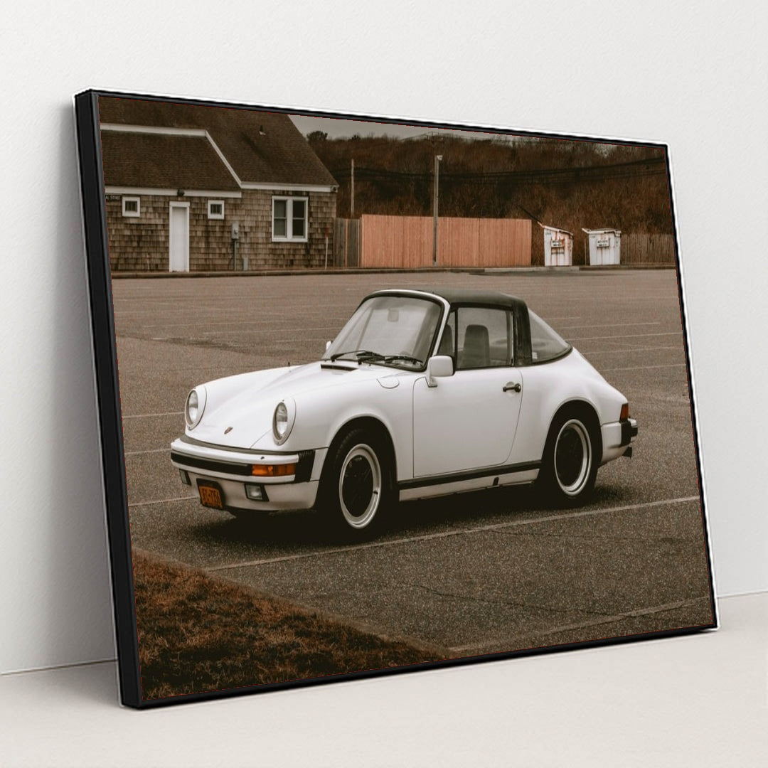 This photo shows a white classic Porsche 911 Targa parked in an empty lot on an overcast day, in a black frame.