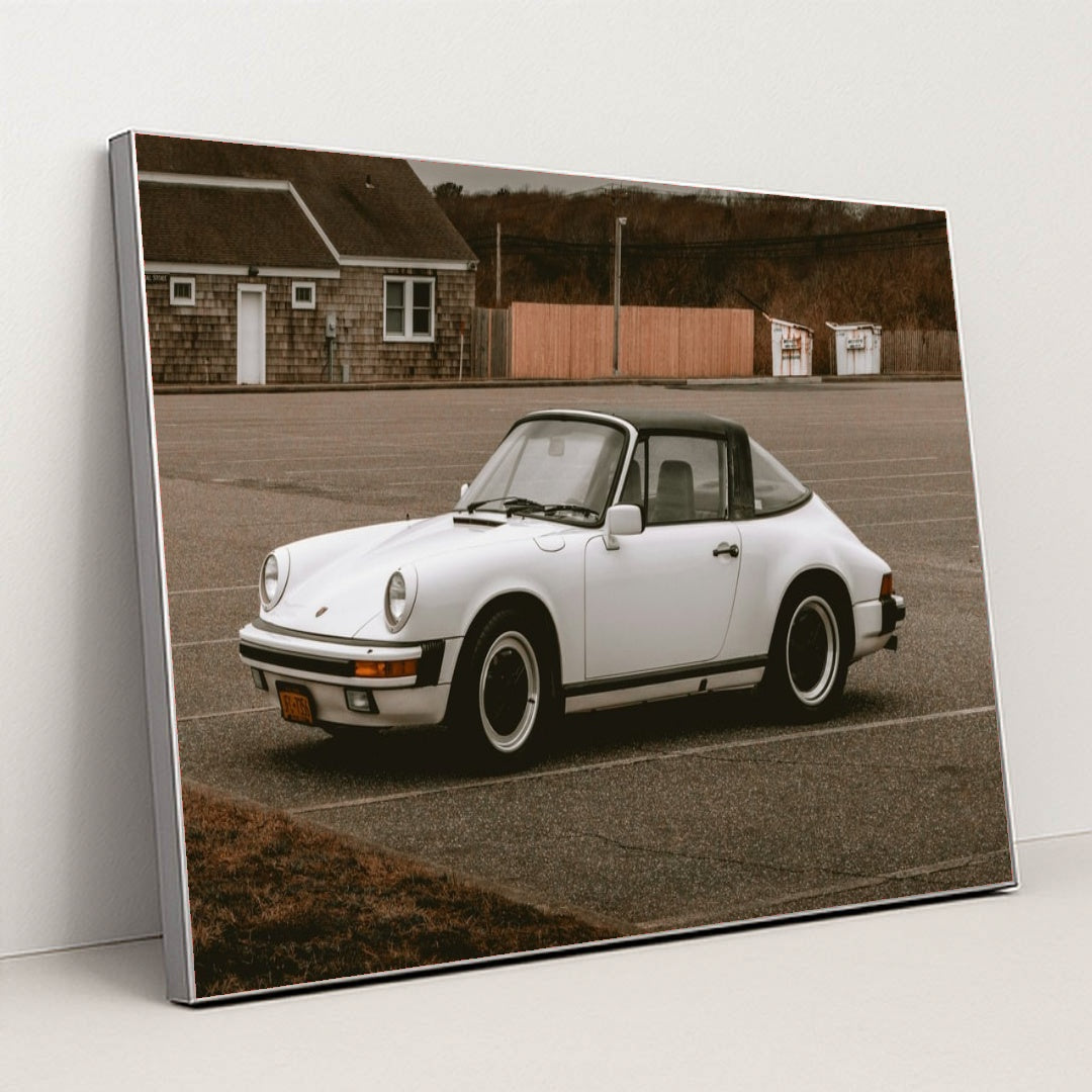 This photo shows a white classic Porsche 911 Targa parked in an empty lot on an overcast day, in a silver frame.