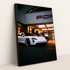 This photo shows a white Porsche Taycan electric sedan parked in front of a Porsche dealership at night, in a black frame.