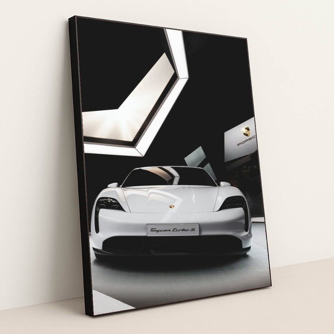 This photo shows the front view of a white Porsche Taycan Turbo S on display in a modern showroom, in a black frame.