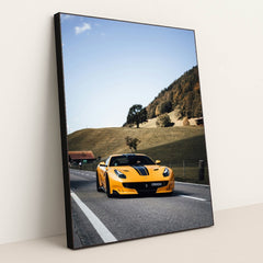 This photo shows a yellow Ferrari sports car with black stripes driving on a rural road by green hills and cottages, in a black frame.