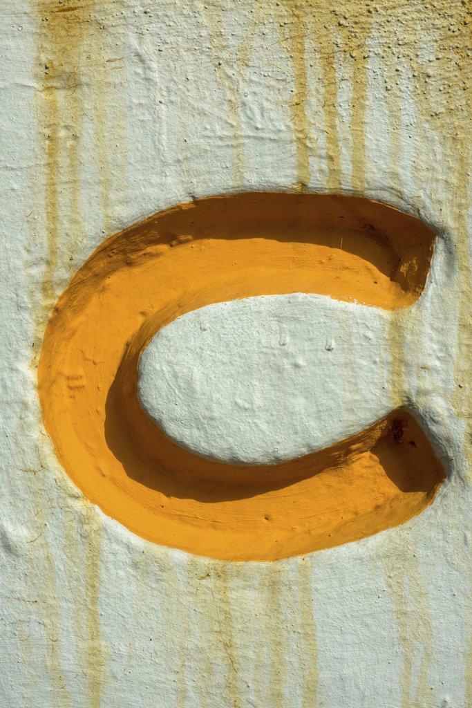 Victor  Moragriega - Close-up of an orange letter C painted on a textured wall with rust drip marks.