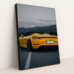 This photo shows the rear of a yellow Porsche 718 Spyder convertible parked on a road with mountains in the background, in a black frame.