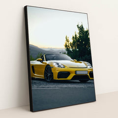 This photo shows a yellow Porsche convertible sports car parked at sunset near green trees and mountains, in a black frame.